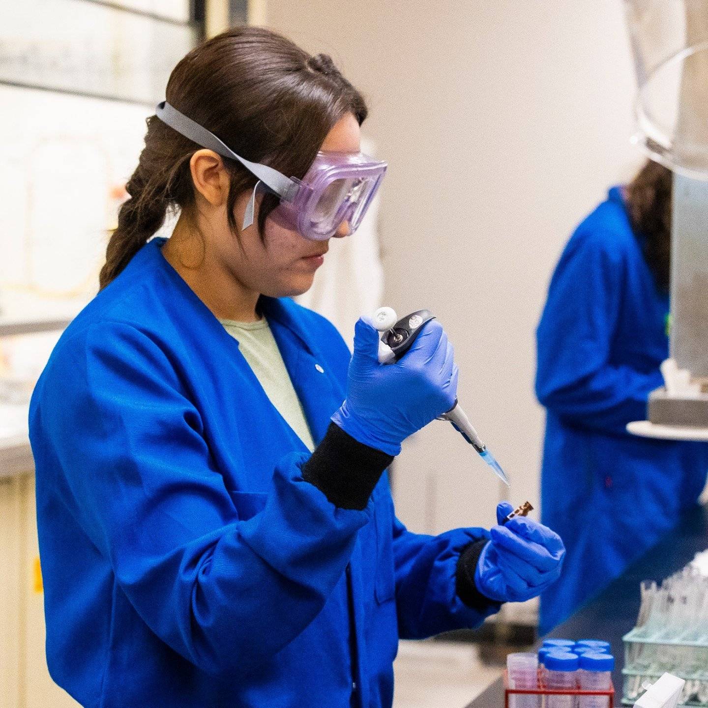 a person wearing goggles and blue coat holding a pipette