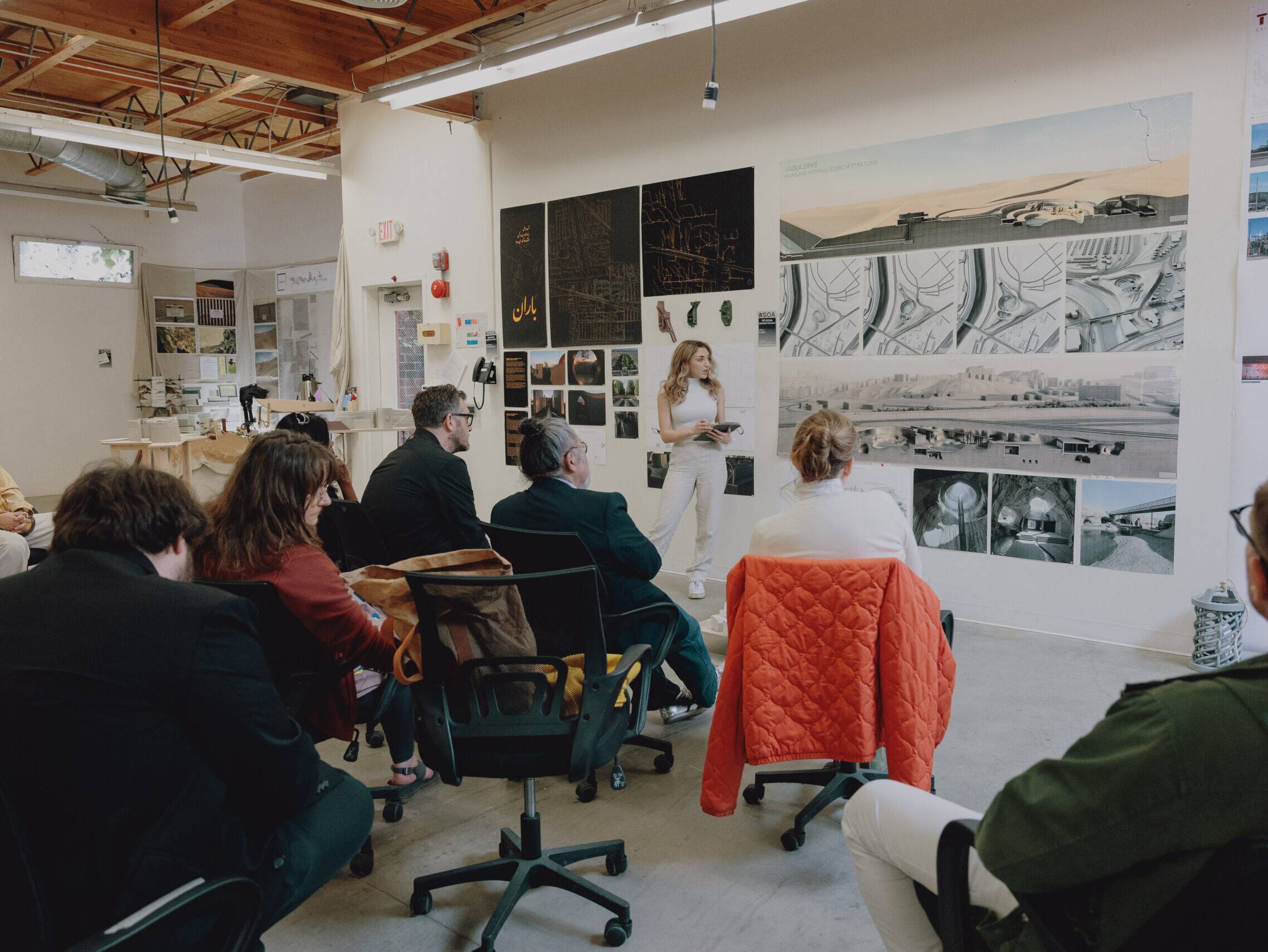 a group of people sitting in chairs in a room with a person standing in front of a white wall