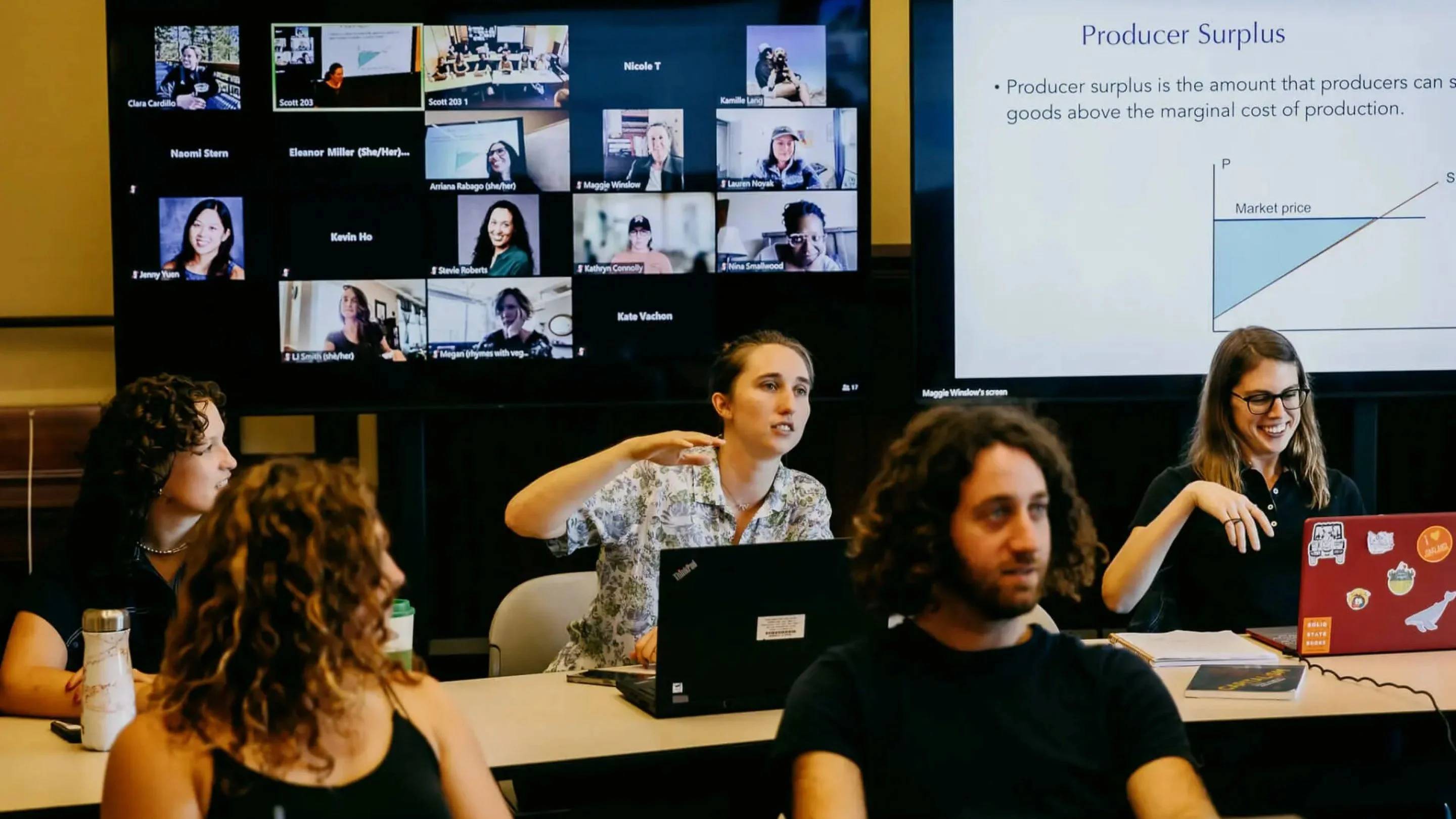 a group of people sitting at a table with laptops and a screen with a presentation