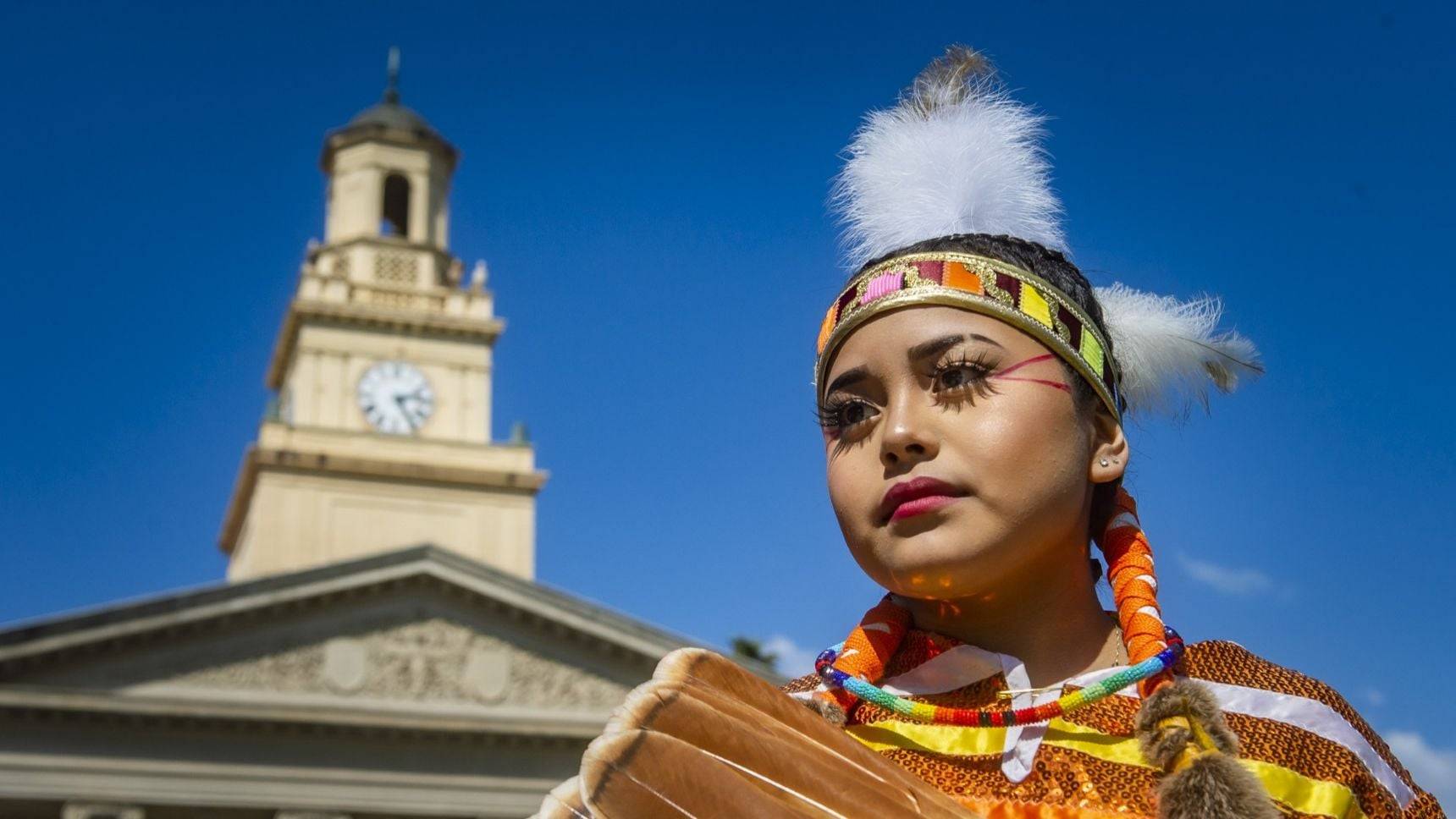 A powwow participant wearing traditional regalia poses in front of Memorial Chapel at University of Redlands.