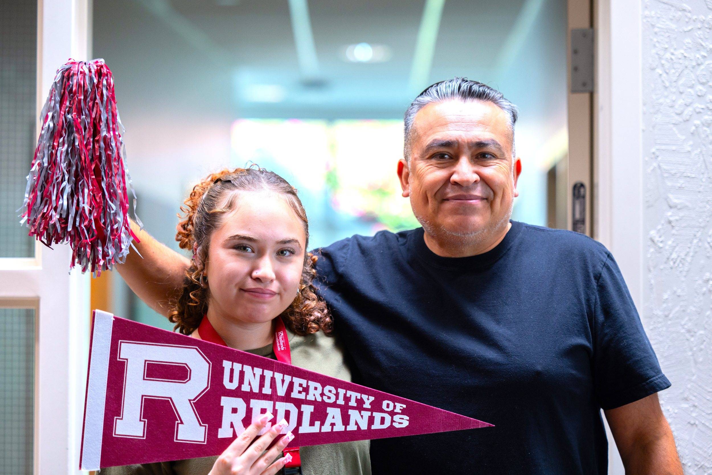 Two people pose with a University of Redlands pennant and a maroon-and-white pom-pom.