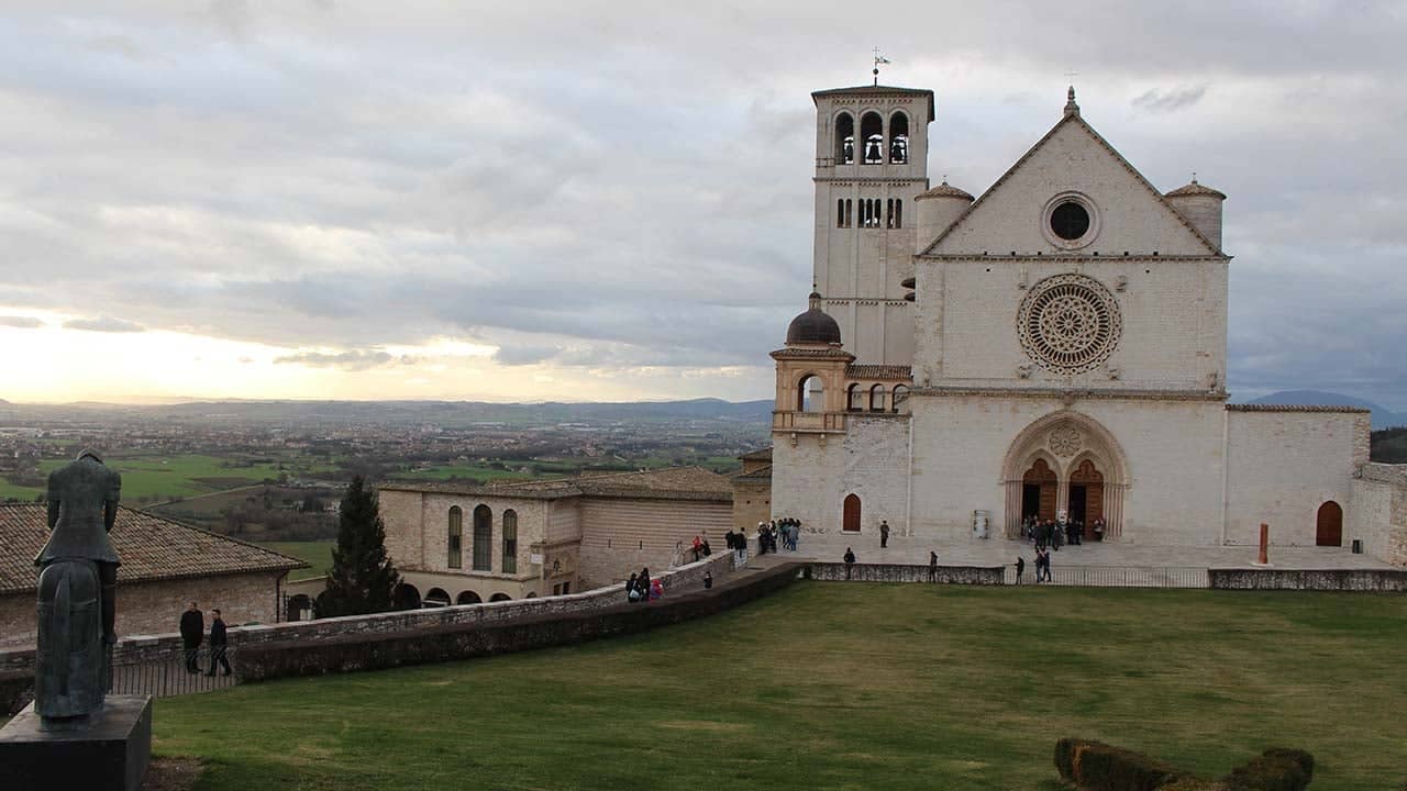 a large white building with a bell tower and a green lawn with Assisi in the background