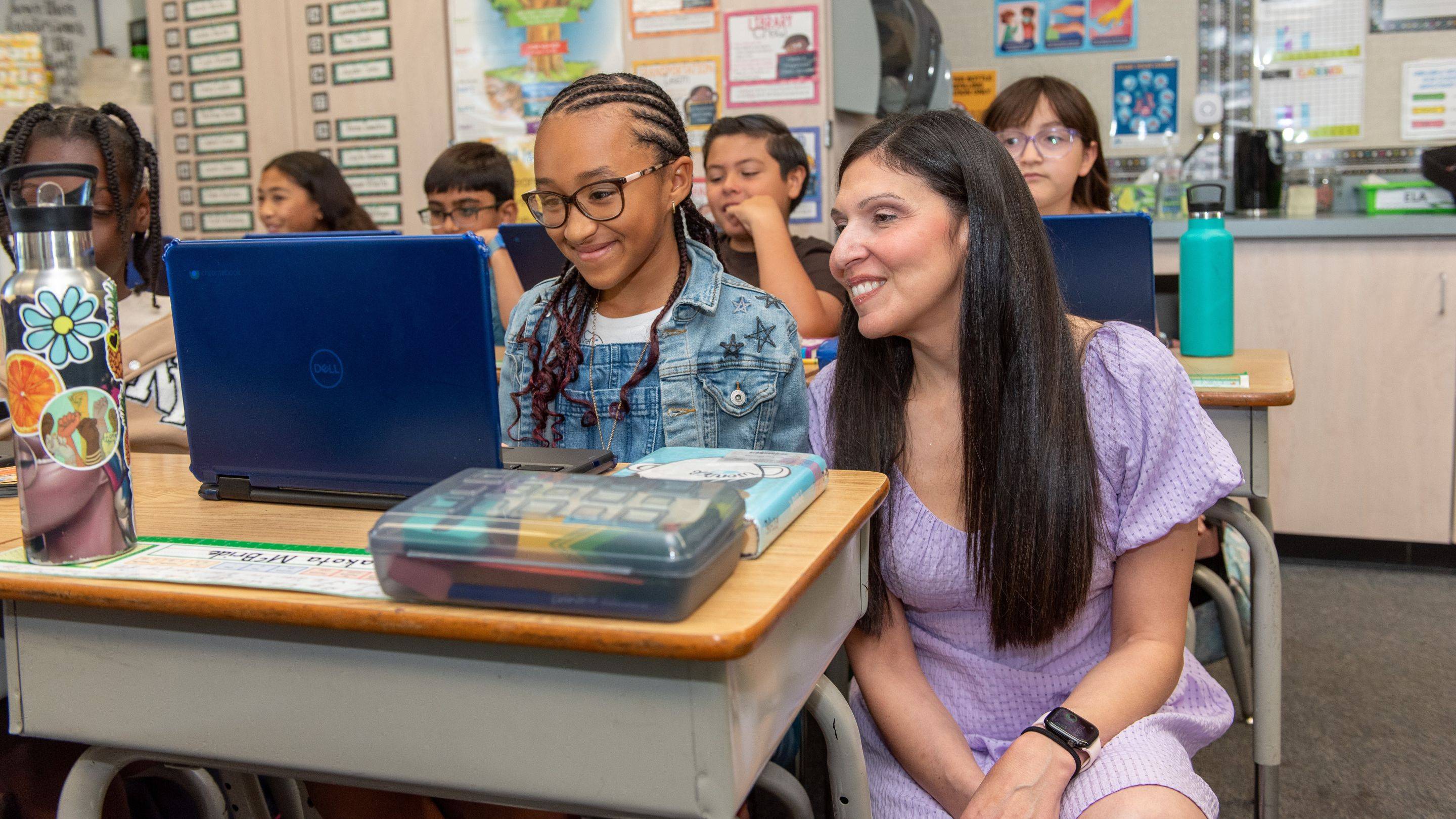 Elementary school teacher helping a student use a laptop at a classroom desk.