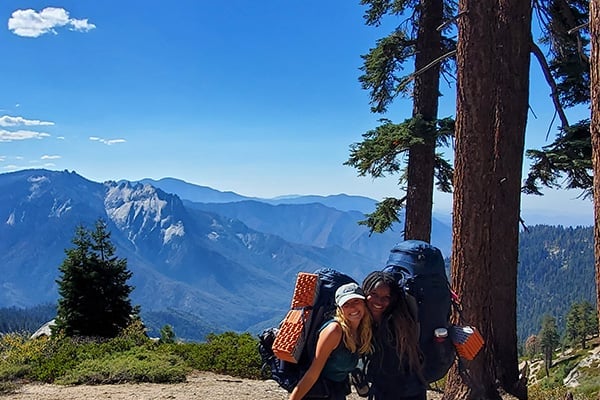 two women standing next to a tree with backpacks