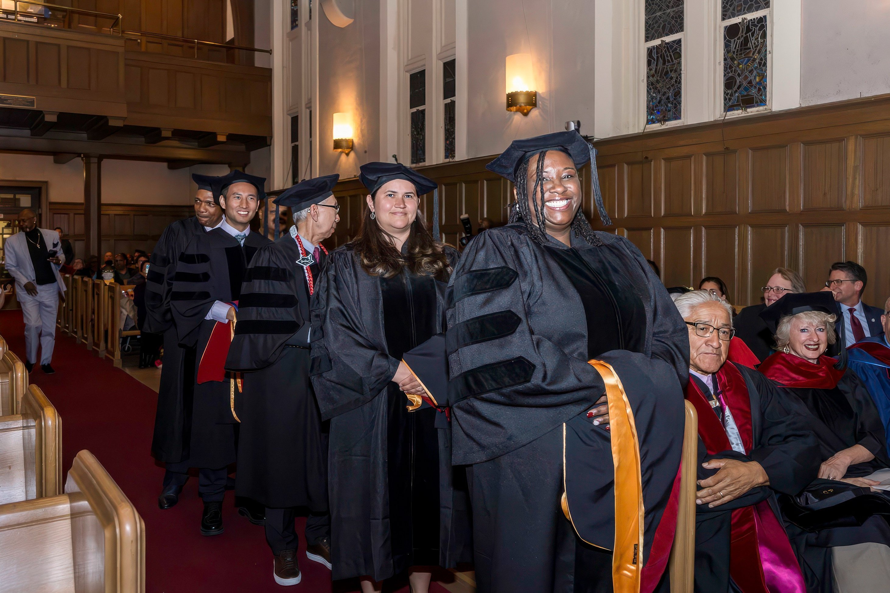 Smiling students wearing black caps and gowns walk down to graduation.