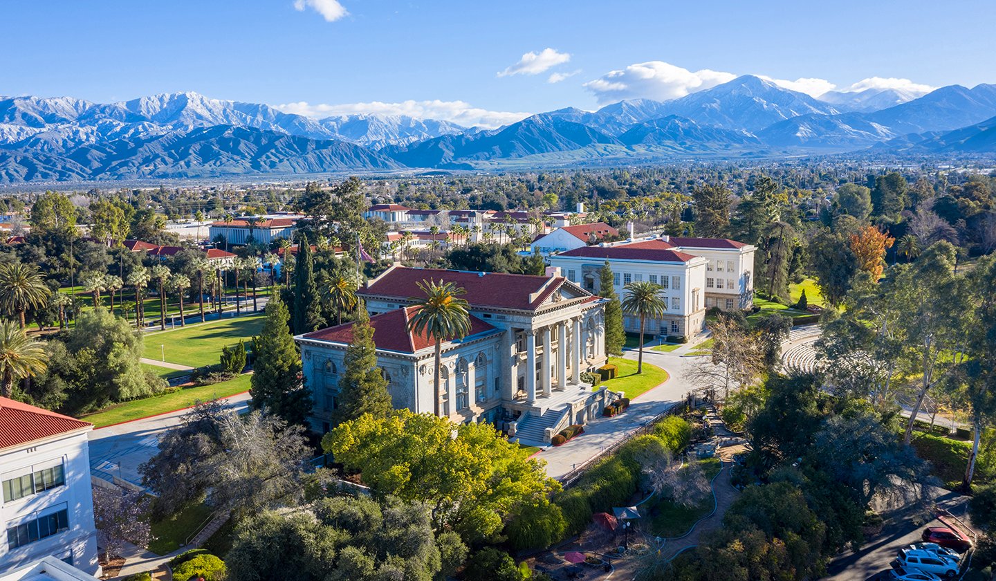 a large white building with a red roof surrounded by trees and mountains