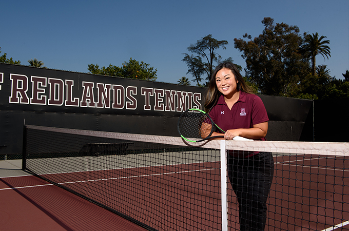 Lauren Villanueva leans against a net on the tennis court, holding a racquet in her hand.