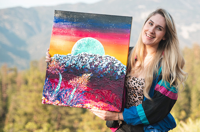Ashley smiles and holds a piece of her artwork up for display.