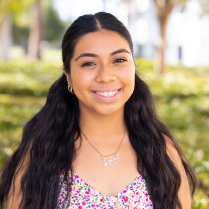 Jennifer Sacor Velasco smiling outdoors in a floral top with greenery in the background.