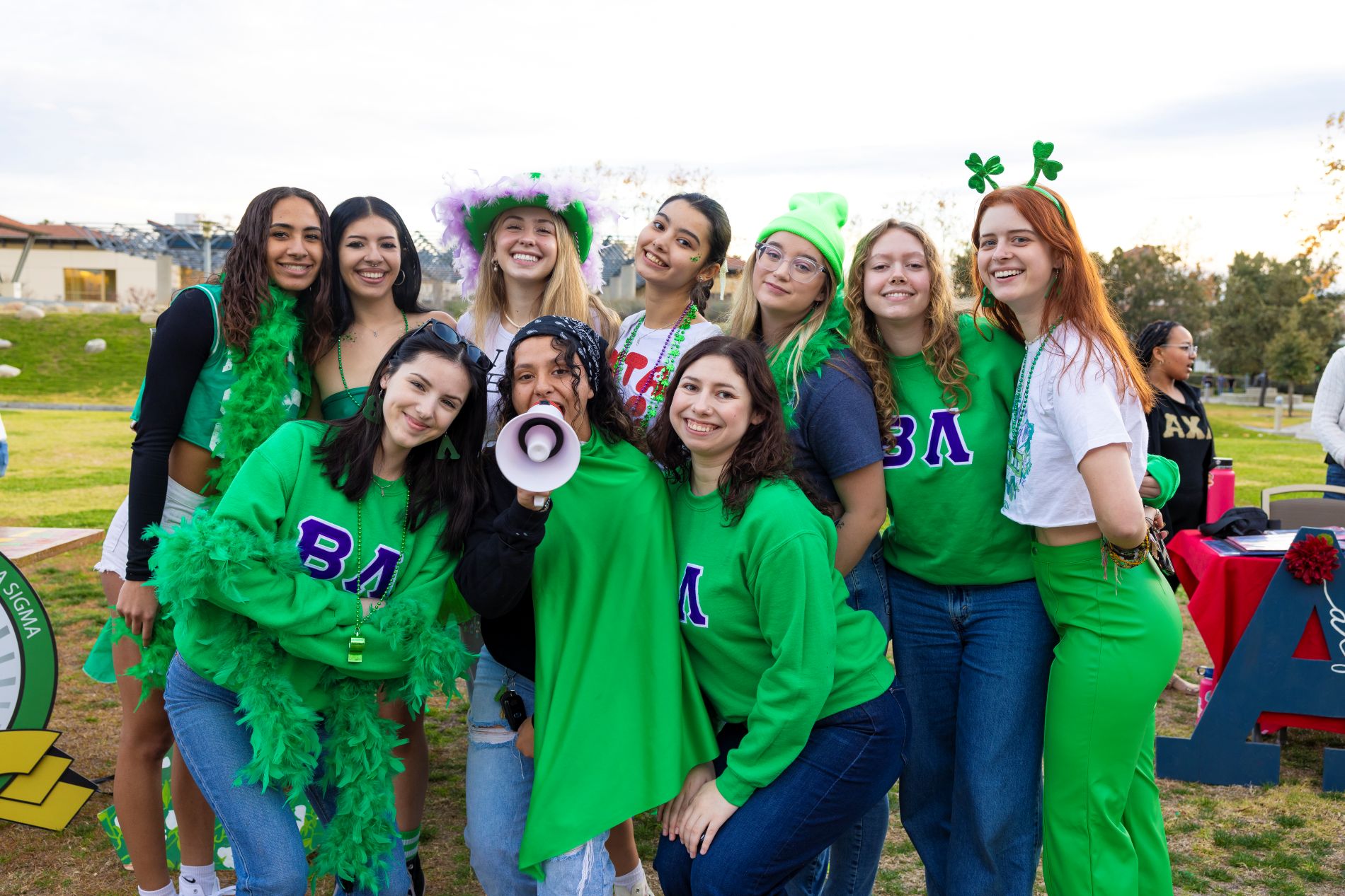 A group of students dressed in green pose together at an outdoor involvement fair event, some wearing festive accessories and sorority letters.