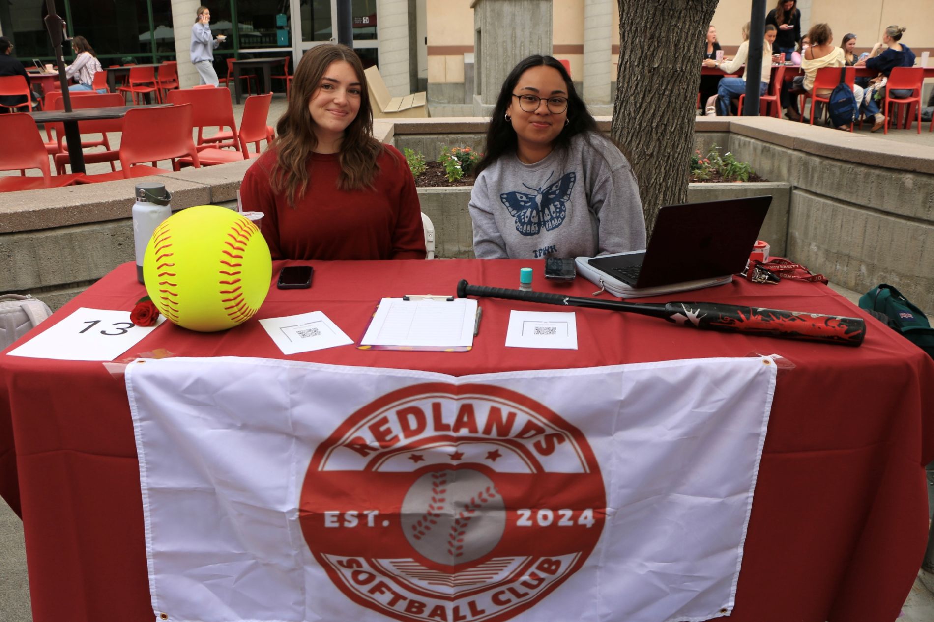 Two students sit behind the Redlands Softball Club booth, with a large decorative softball, a softball bat, and a club banner on display.